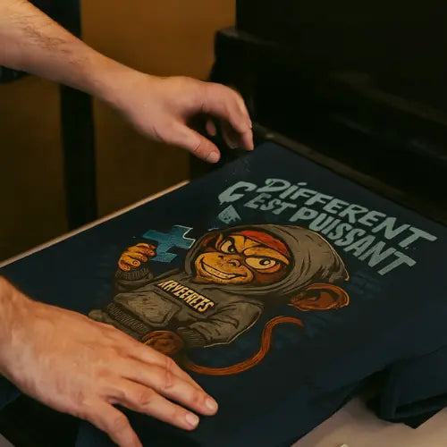 Un t-shirt Captain Cawette fraîchement imprimé avec le message "Différent c’est puissant", posé sur un plan de travail, avec une étiquette indiquant "Printé pour : Noemy". Impression textile personnalisée en print-on-demand