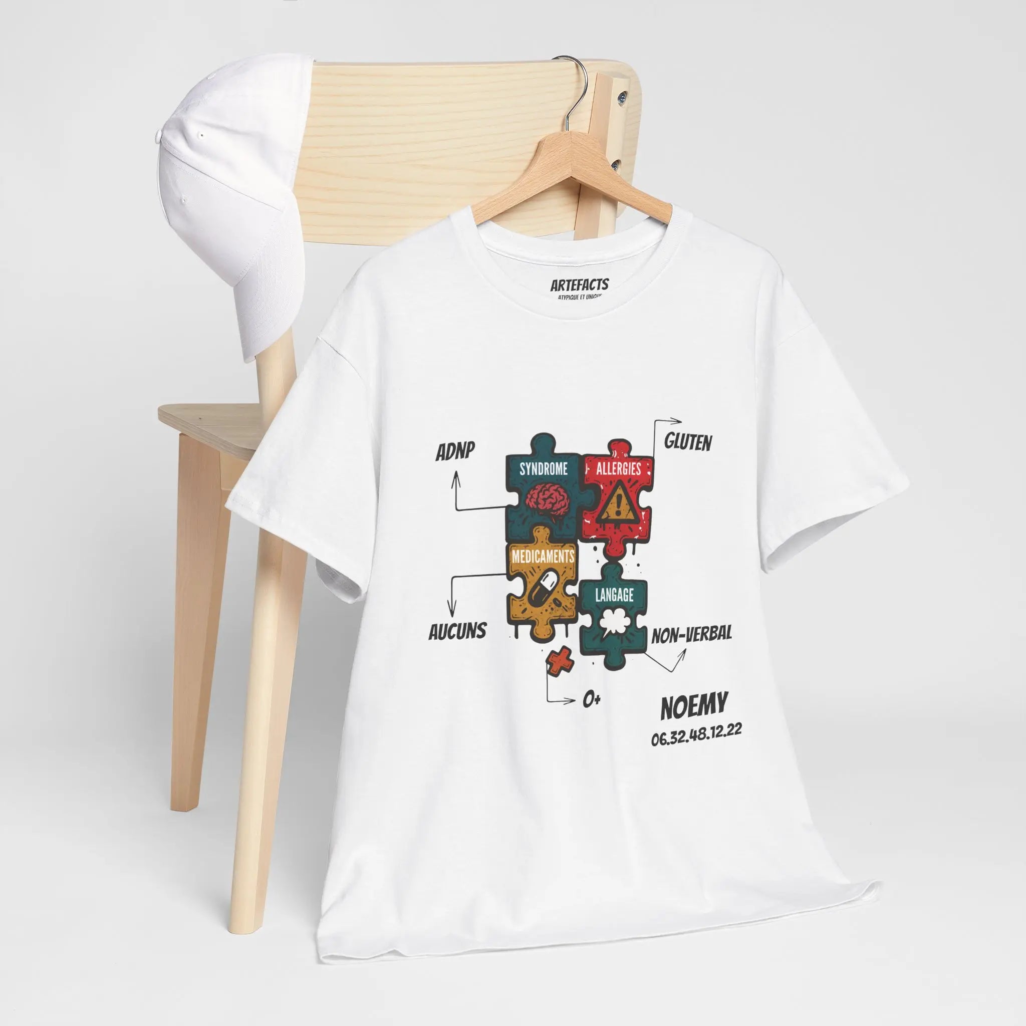 T-shirt blanc personnalisé accroché à une chaise, affichant des pièces de puzzle colorées représentant les informations médicales de Noemy : syndrome ADNP, allergies, médicaments, langage, groupe sanguin et autres éléments essentiels.
